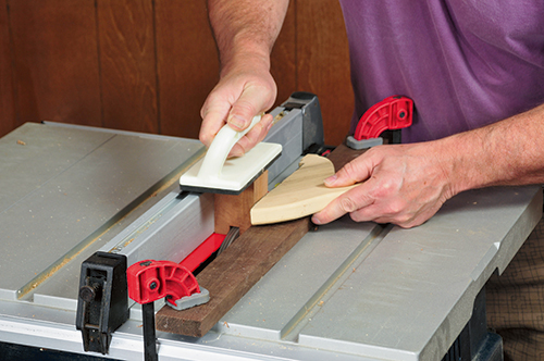 A 1/2"-wide dado blade cuts a groove in the uke ornament’s body blank for the neck. Clamping a rail to the table saw top helps keep the small workpiece aligned during cutting.