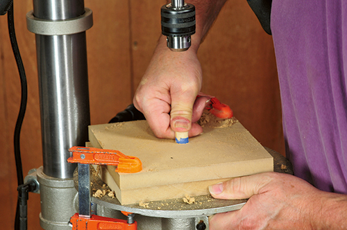 A 5/8"-dia. dowel, which will become the uke ornament’s sound hole ring, is pressed into a hole bored partway through two pieces of MDF or plywood clamped atop the drill press table.